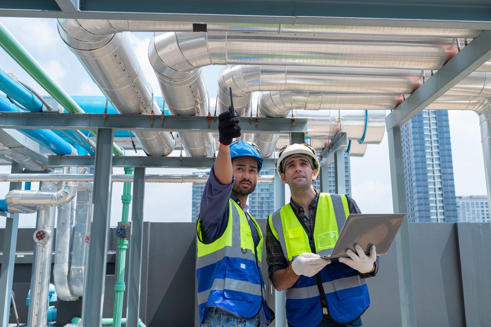 Two engineers in safety vests and helmets collaborate and share a laugh while working on industrial machinery in a facility.