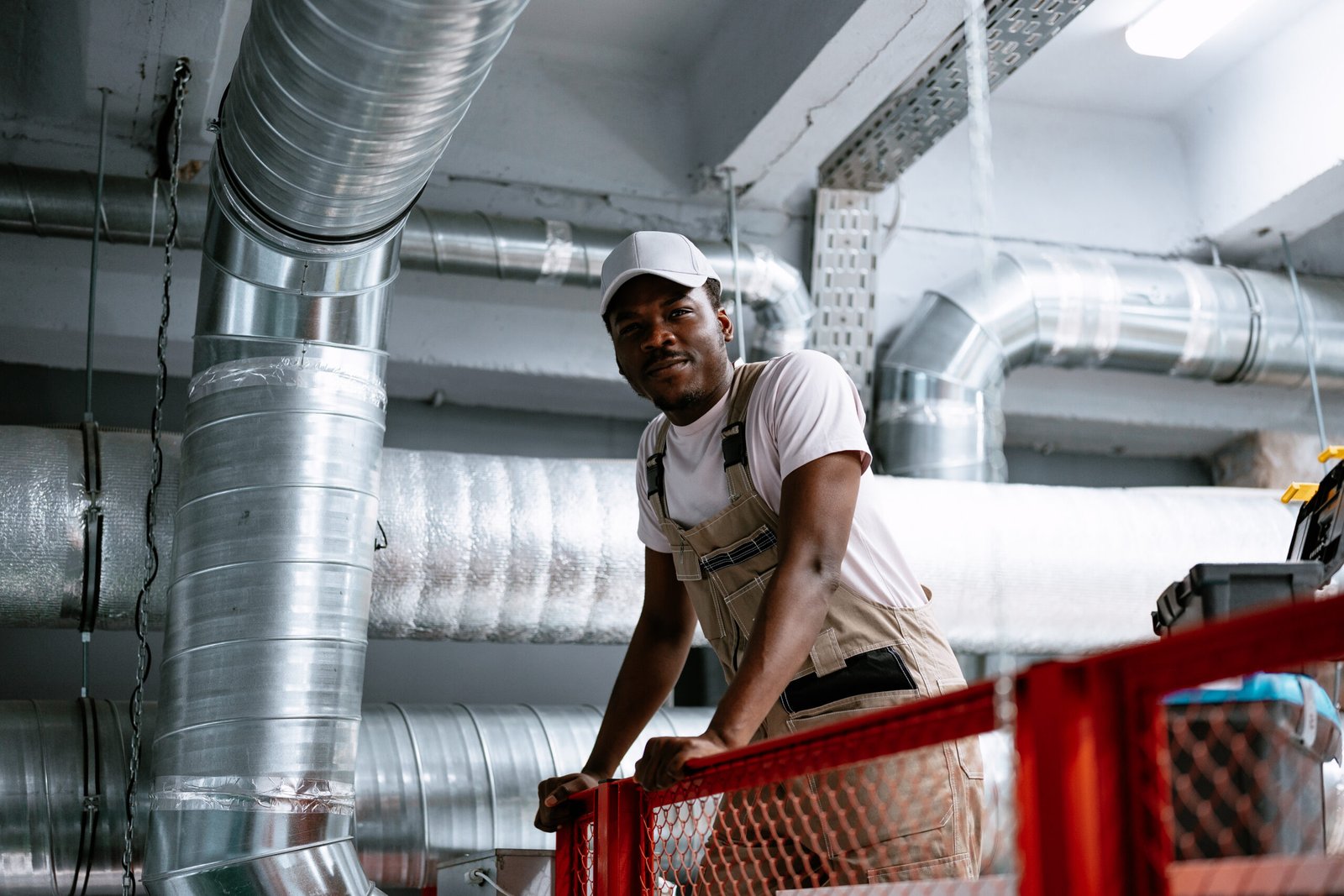 A worker is positioned on a raised platform, checking the ventilation ducts in an industrial setting. Bright lights illuminate the area as he focuses on his task.